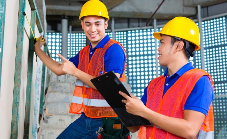 建設現場で図面を確認しながらメジャーで計測作業を行うヘルメット姿の2人の作業員