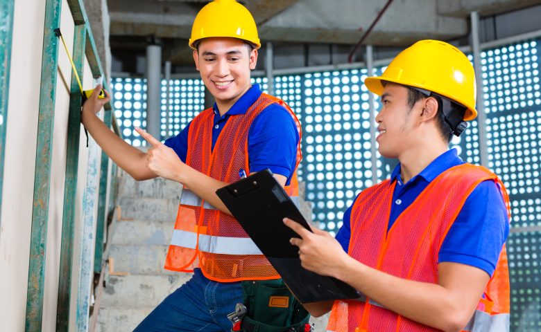 建設現場で図面を確認しながらメジャーで計測作業を行うヘルメット姿の2人の作業員