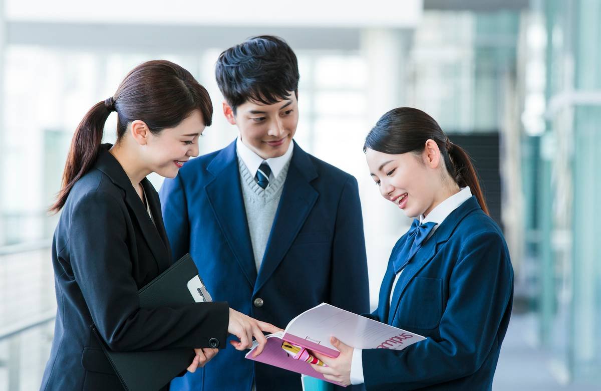 塾のパンフレットを見ながら先生に進路相談をする制服姿の高校生男女と大学受験の対策イメージ